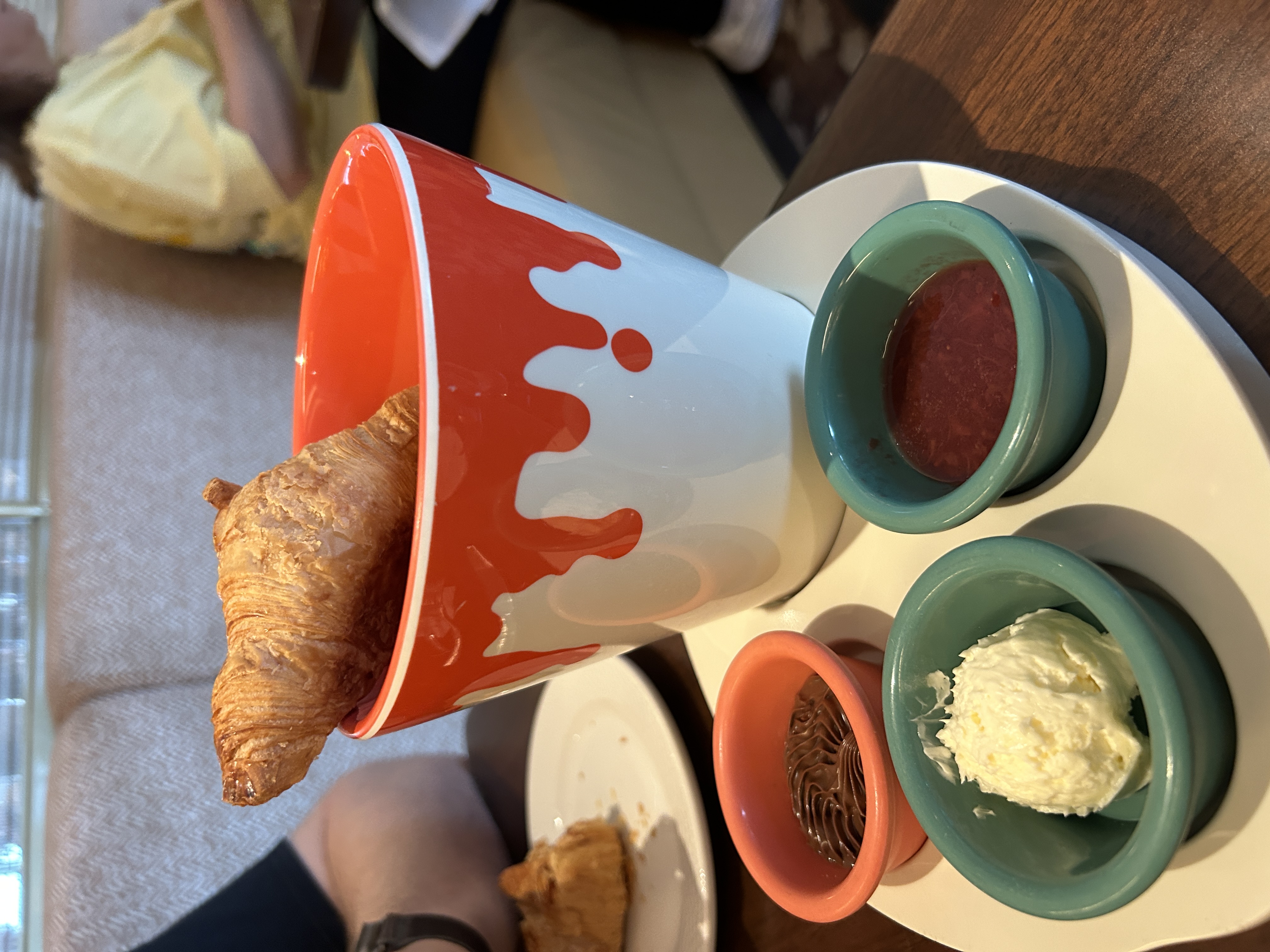 Image shows a ceramic pot with a croissant in and three pots of jams the pot is white with what is supposed to be red pain as an effect