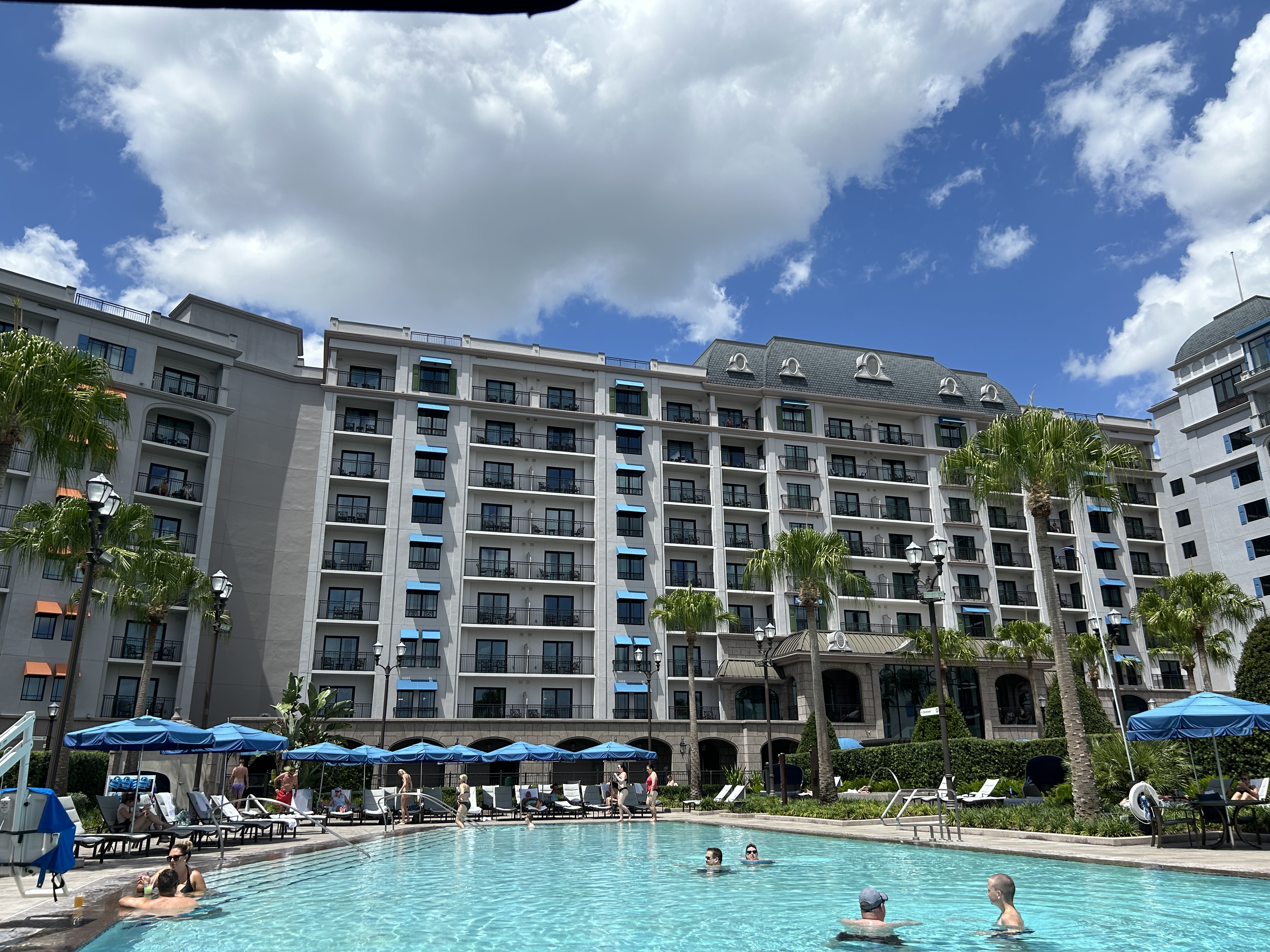Image shows a swimming pool with a hotel building behind and blue sky